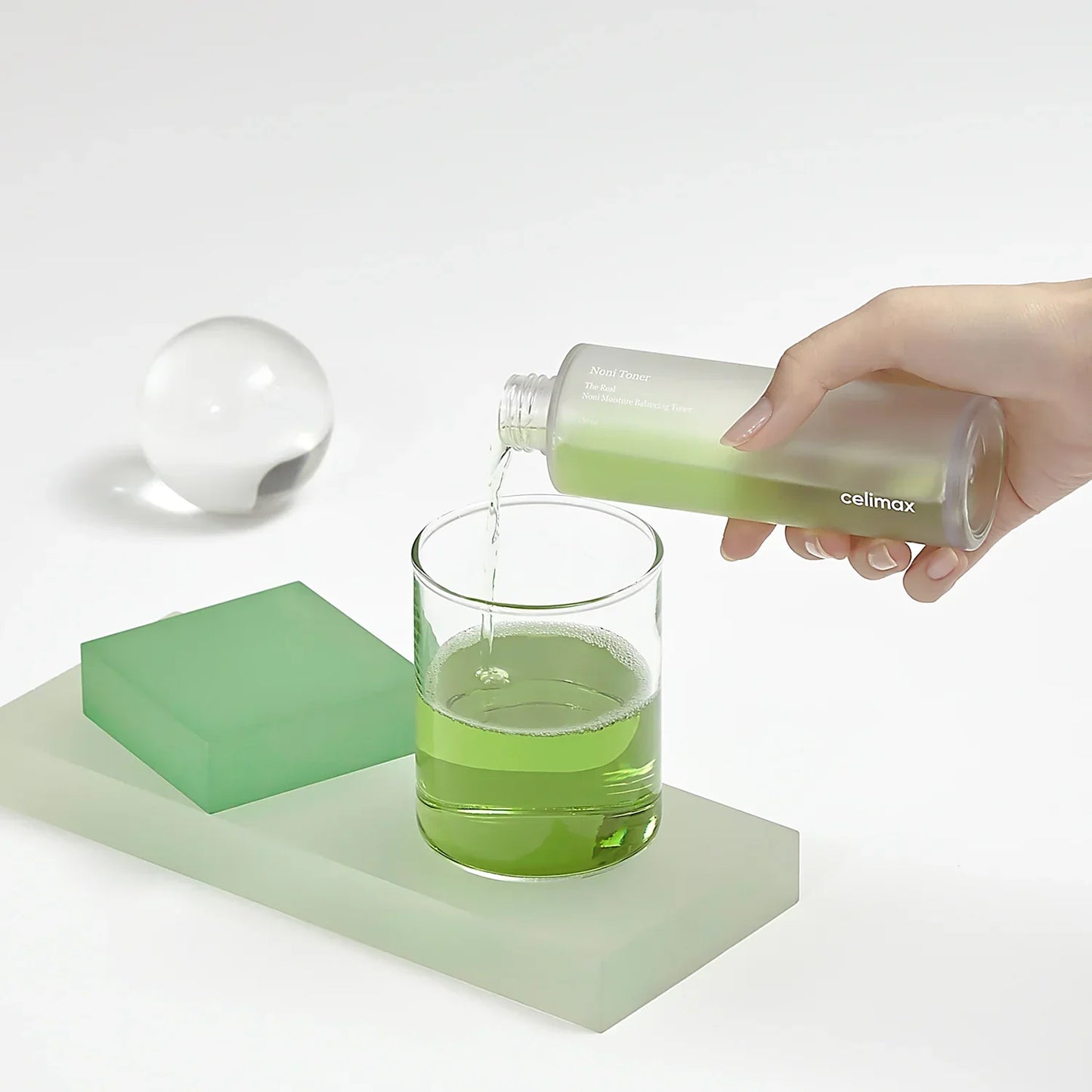 Tónico Noni verde siendo vertido en vaso de cristal sobre base blanca con objetos decorativos verdes
